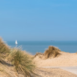 Zandduinen en zeezicht nabij Westende, België, met helmgras en een zeilboot aan de horizon.