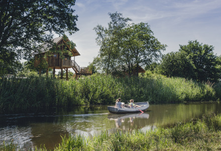 Twee mensen varen in een roeiboot op een rivier met een boomhut bij vakantiepark Mölke in Overijssel.