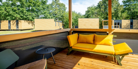 Eine Terrasse mit gelbem Sofa im Urban-Gardens Ferienpark in Ostflandern, Belgien, umgeben von Holzhütten.