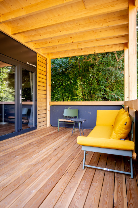 Covered wooden terrace with yellow sofa, chair, and forest view at Urban-Gardens holiday park, Belgium.