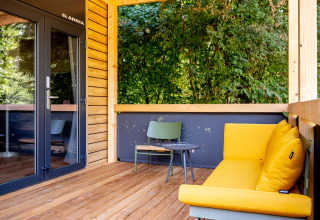 Überdachte Holzterrasse mit gelbem Sofa, Stuhl und Waldblick im Urban-Gardens Ferienpark, Ostflandern.