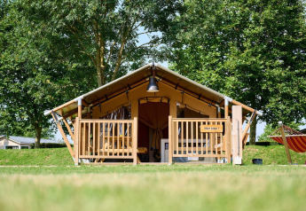 Tenda safari con veranda in legno e amaca sul prato, circondata da alberi verdi in una giornata di sole.