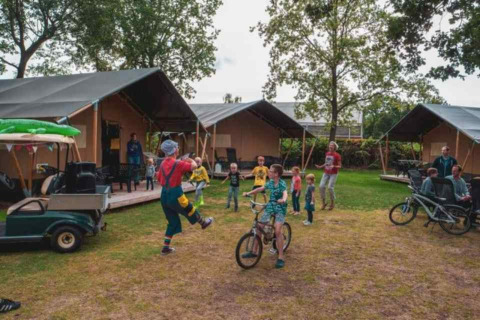 Niños juegan y andan en bicicleta frente a tiendas safari en Camping 't Veld, Países Bajos, con familias alrededor.