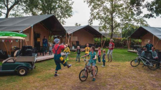 Niños juegan y andan en bicicleta frente a tiendas safari en Camping 't Veld, Países Bajos, con familias alrededor.