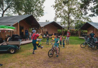 Niños juegan y andan en bicicleta frente a tiendas safari en Camping 't Veld, Países Bajos, con familias alrededor.