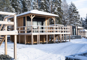Chalets en bois du lodge Edelweiss sous la neige, fenêtres vitrées, terrasse surélevée et forêt derrière.
