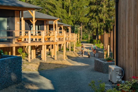 Familie fährt Fahrrad vor Holzbungalows der Edelweiss Lodge, umgeben von Bäumen und Grünpflanzen.