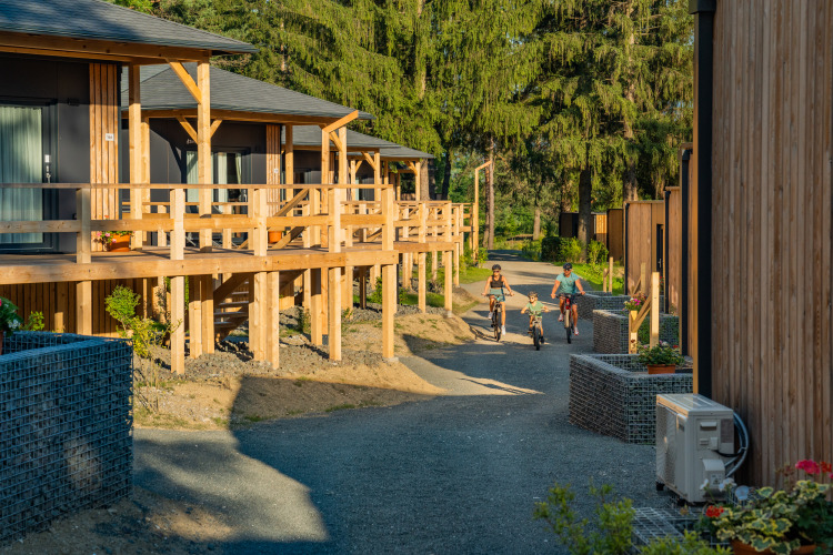 Gezin fietst bij houten lodges van Edelweiss Lodge, omgeven door bomen en bloeiende planten.