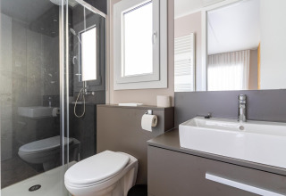 Baño moderno en la cabaña Edelweiss con ducha de vidrio, inodoro, lavabo y espejo grande iluminado.