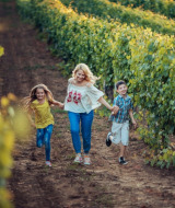 A woman and two children run joyfully through lush vineyards near Sigean, Occitanie, France, surrounded by grape vines.