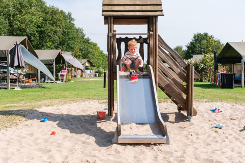 Niño jugando en un tobogán en un área de arena rodeado de tiendas en Kampeerdorp de Zandstuve, Overijssel.
