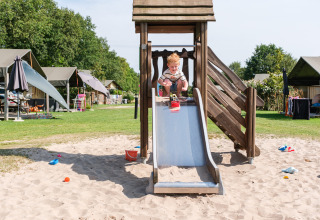 Kind spielt auf einer Rutsche im Sandgebiet, umgeben von Zelten im Ferienpark Kampeerdorp de Zandstuve.