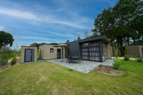 Moderne L-Cube 4+2 lodge met terras, tuinmeubilair en veel groen onder een stralend blauwe lucht.