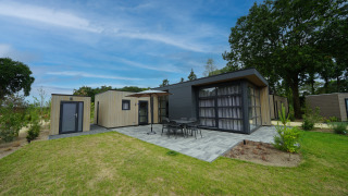 Moderne L-Cube 4+2 lodge met terras, tuinmeubilair en groene tuin onder een heldere blauwe lucht.