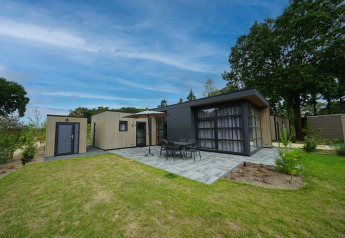 Moderne L-Cube 4+2 lodge met terras, tuinmeubilair en groene tuin onder een heldere blauwe lucht.