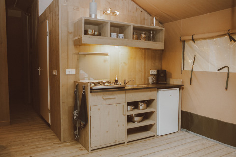 Luxury safari tent kitchenette with wooden decor, gas stove, sink, mini fridge, and open shelves in the Netherlands.