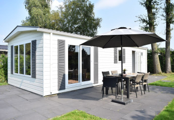 Veluwelodge met witte buitenkant, terras, parasol en tuinmeubilair op Holiday park De Boshoek in Nederland.