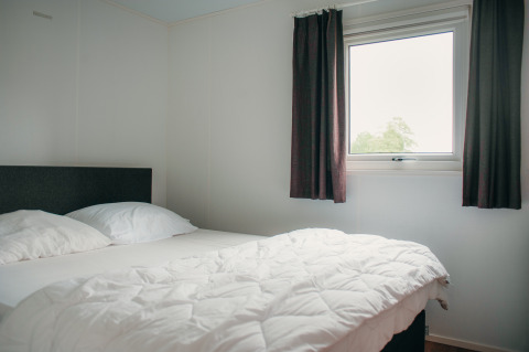 Schlafzimmer in der Veluwelodge im Ferienpark De Boshoek, Niederlande, mit Bett, Fenster und Vorhängen.