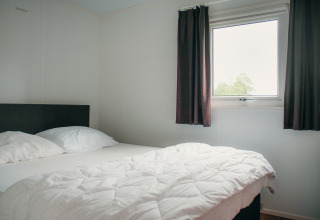 Schlafzimmer in der Veluwelodge im Ferienpark De Boshoek, Niederlande, mit Bett, Fenster und Vorhängen.