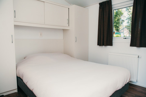 Slaapkamer met dubbel bed, witte kasten, gordijnen en raam in Veluwe lodge + sauna, De Boshoek, Nederland.