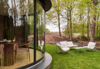 Zona de estar exterior junto a la ventana curva de la tiny house Lumni Pod en Wilsumer Berge, Alemania, rodeada de árboles.