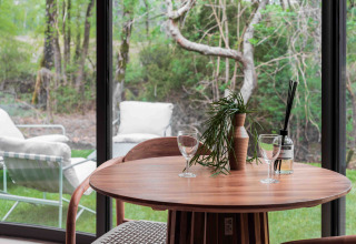 Mesa de madera con dos copas de vino y decoración junto a ventanales con vistas verdes en Lumni Pod, Wilsumer Berge.