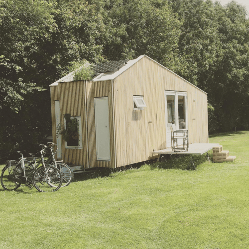 Petit chalet en bois du Het Eilandhuis sur pelouse avec vélos et terrasse, entouré d’arbres verdoyants.