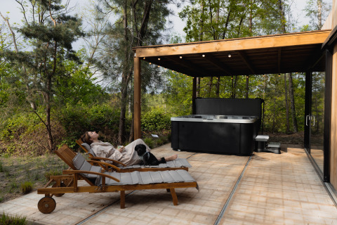 Donna rilassata con il suo cane su una sdraio vicino alla vasca idromassaggio a Comfort Sweet Wellness, Cosy Cabins, Limburgo, Belgio.