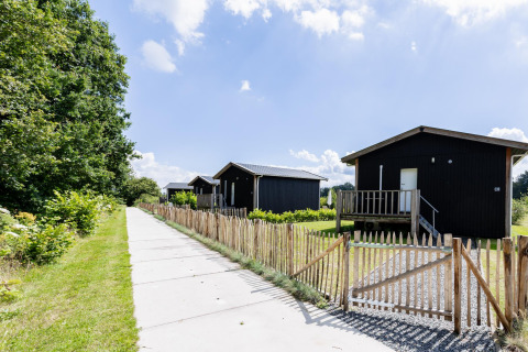 Tiny House XL bei The Kremmer in den Niederlanden zeigt schwarze Holzchalets neben einem Weg im Grünen.