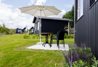 Außenbereich des Tiny House XL bei The Kremmer, Niederlande mit schwarzen Häusern, Stühlen und Sonnenschirm.
