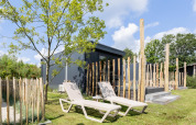 Due lettini prendisole su prato davanti a una tiny house con terrazza, circondata da pali di legno e alberi.