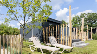Due lettini prendisole su prato davanti a una tiny house con terrazza, circondata da pali di legno e alberi.