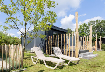 Due lettini prendisole su prato davanti a una tiny house con terrazza, circondata da pali di legno e alberi.
