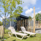 To liggestole står på græsset foran en tiny house med terrasse, træer og træhegn udendørs om sommeren.