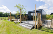 Modern tiny house with deck and wood fencing, sunny day at The Kremmer in the Netherlands, lush lawn.