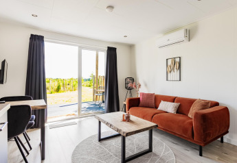Lyst tiny house med brun sofa, træbord og udsigt til have via glasskydedøre på The Kremmer, Nederlandene.