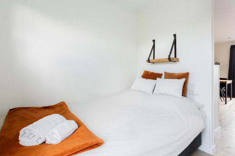 Dormitorio luminoso en tiny house con cama doble, manta naranja y toallas en The Kremmer, Países Bajos.
