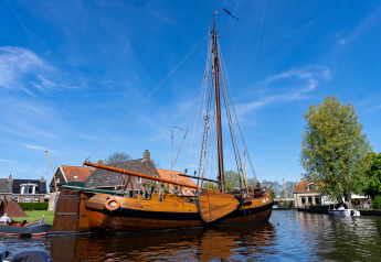 Traditional wooden sailboat docked at Marina Heeg in the Netherlands, with buildings and trees nearby.