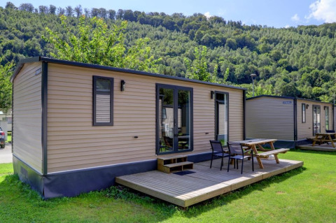 A photo of modern cabins with wooden decks and picnic tables, set on green grass near forested hills.
