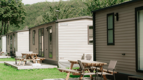 Fila de cabañas modernas con terrazas y mesas de picnic en un entorno verde y arbolado durante el verano.