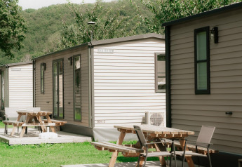 Fila de cabañas modernas con terrazas y mesas de picnic en un entorno verde y arbolado durante el verano.