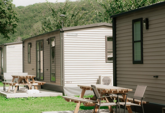 Reihe moderner Hütten mit Terrassen und Picknicktischen in einer grünen, bewaldeten Sommerlandschaft.
