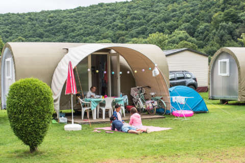Famiglia rilassata davanti a una tenda safari Coco Sweet in campeggio, con biciclette e colline sullo sfondo.