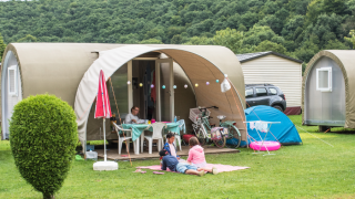 Family relaxing in front of a Coco Sweet safari tent on a campsite, with bikes, tent, and hills behind.