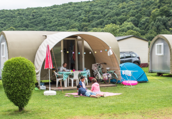 Famiglia rilassata davanti a una tenda safari Coco Sweet in campeggio, con biciclette e colline sullo sfondo.