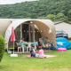 Famiglia rilassata davanti a una tenda safari Coco Sweet in campeggio, con biciclette e colline sullo sfondo.
