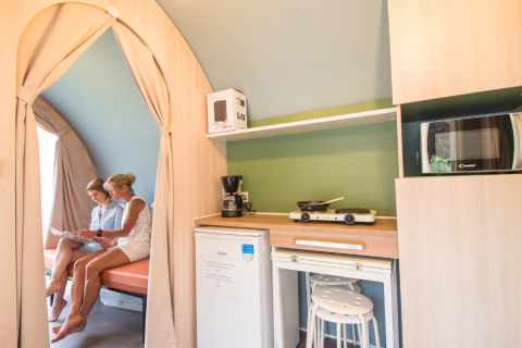 Twee vrouwen zitten in een Coco Sweet safaritent met kitchenette, koelkast en magnetron.