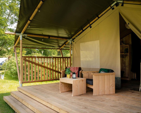 Sistemazione glamping con terrazza in legno coperta, area relax e arredi esterni immersi nel verde.