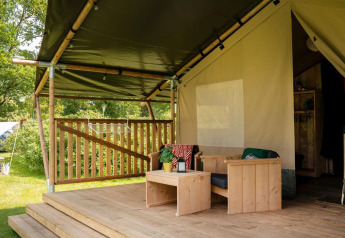 Sistemazione glamping con terrazza in legno coperta, area relax e arredi esterni immersi nel verde.