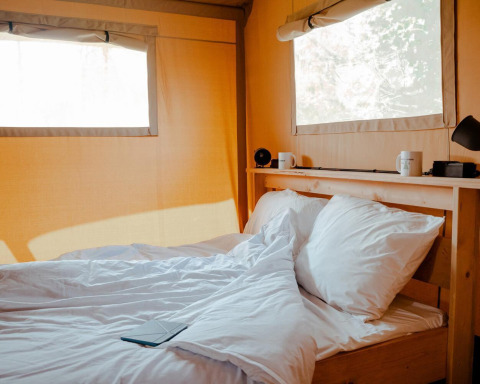 Cama acogedora con sábanas blancas en una tienda de glamping iluminada y ventanas grandes de lona clara.
